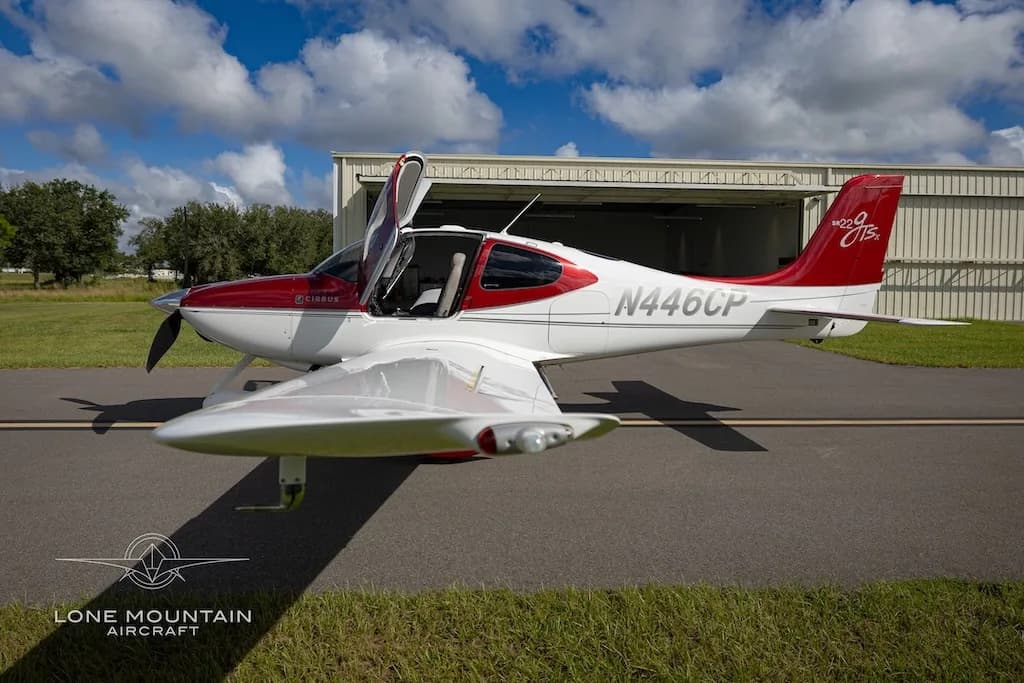 A red and white Cirrus SR22 aircraft (N446CP) with its doors open, viewed from the front-right, parked on an asphalt ramp.