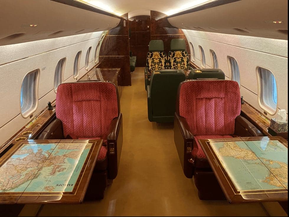 The main cabin of a Bombardier Challenger 604, featuring red velvet seats, dark wood tables with map inlays, and patterned seats in the background.