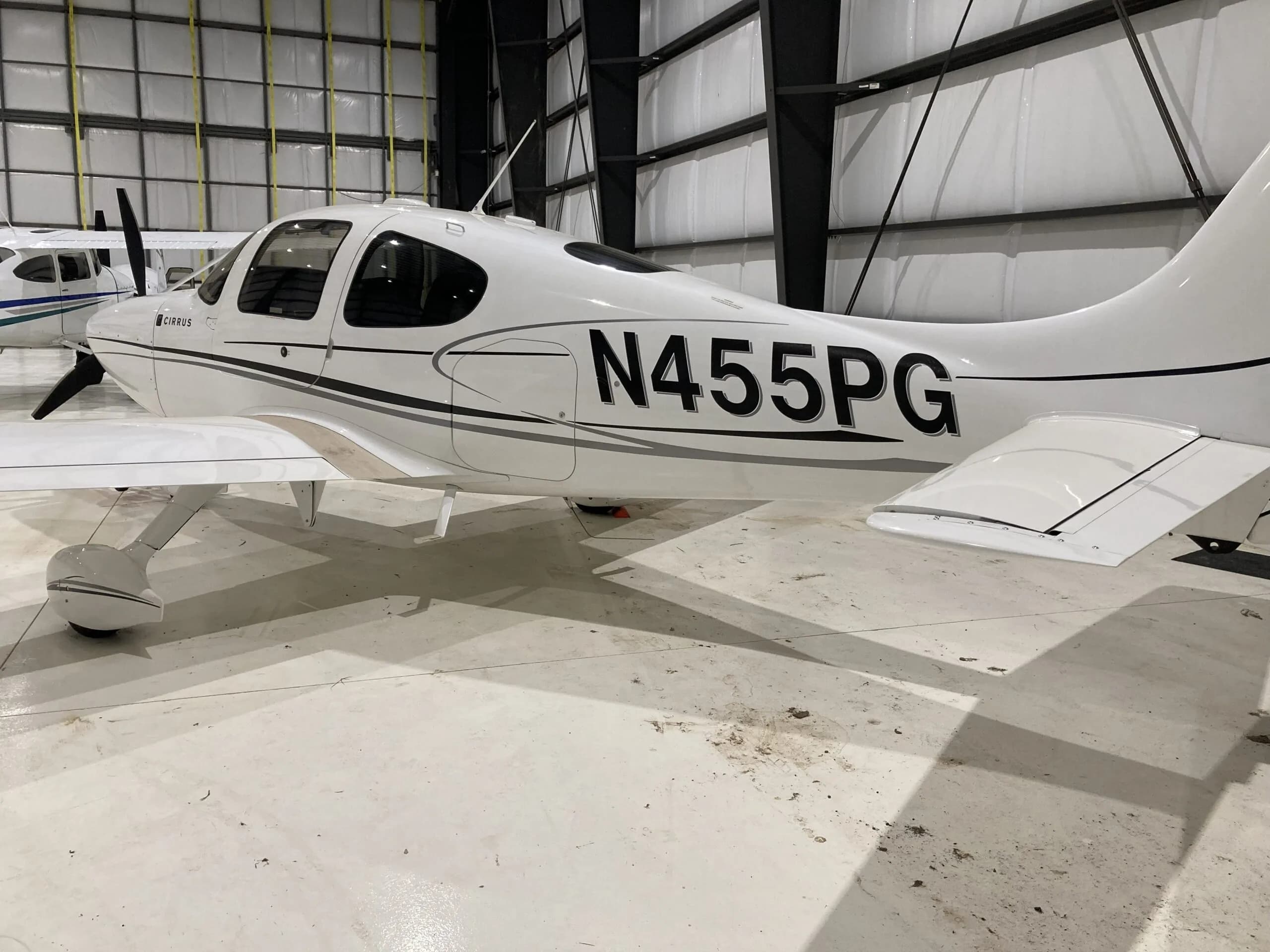 Rear-side view of a white Cirrus SR22 aircraft with black and grey stripes, registration N455PG, in a hangar.