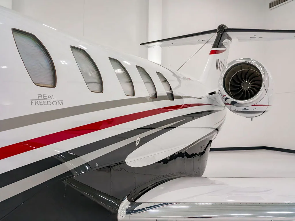 A close-up of the rear fuselage and engine of a white Cessna Citation Latitude, showing black, red, and grey stripes and "REAL FREEDOM" text.