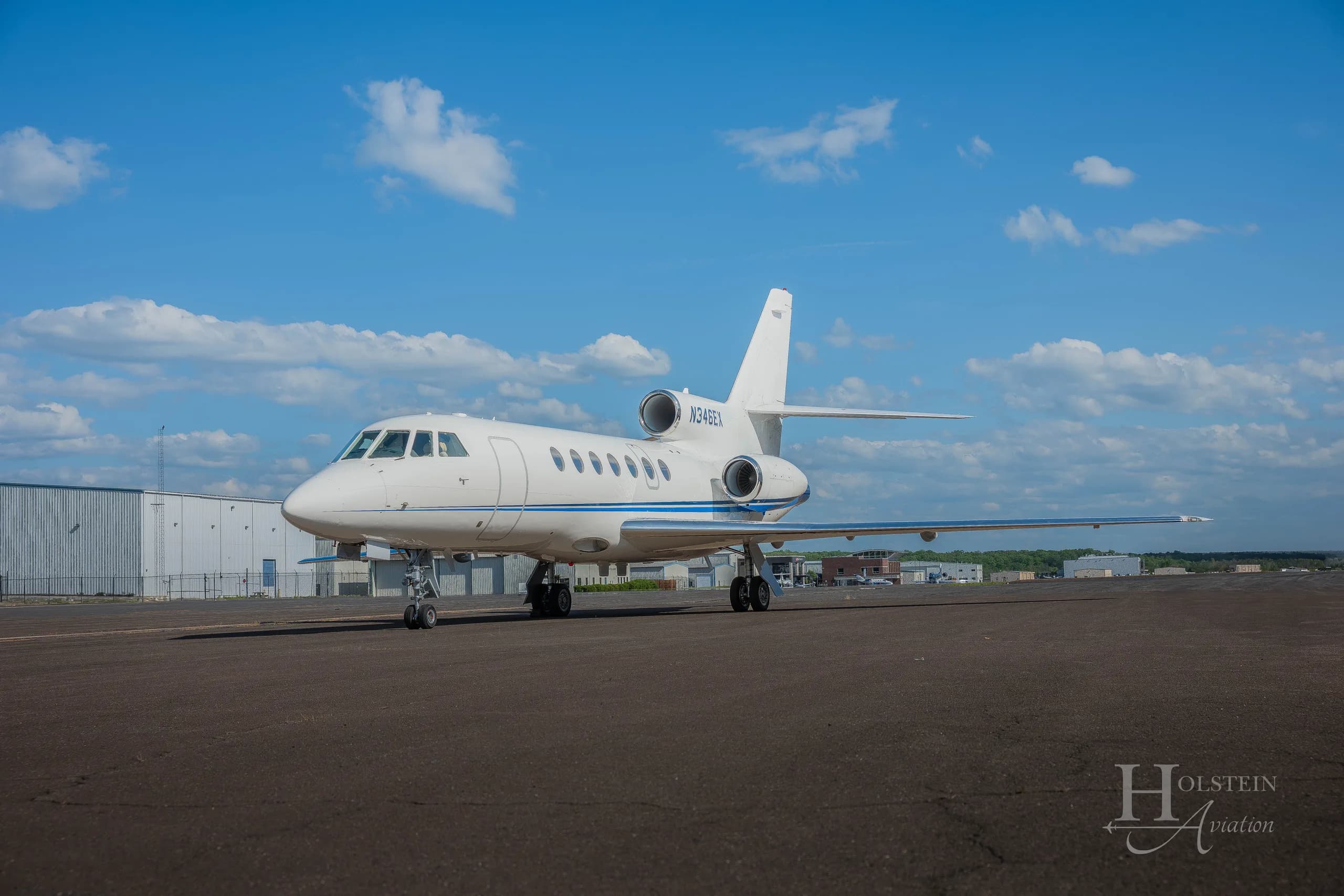 A white Dassault Falcon 900EX business jet with a blue stripe, registration N346EX, parked on an airfield under a blue sky.