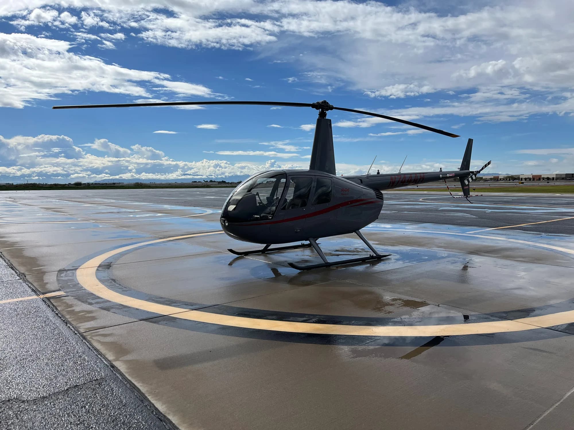 A grey Robinson R44 helicopter parked on a wet tarmac at an airport under a cloudy sky.