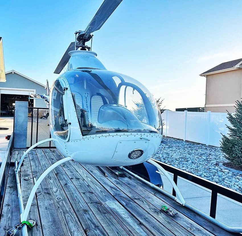 Front view of a white helicopter on a wooden trailer, parked in a gravel driveway.