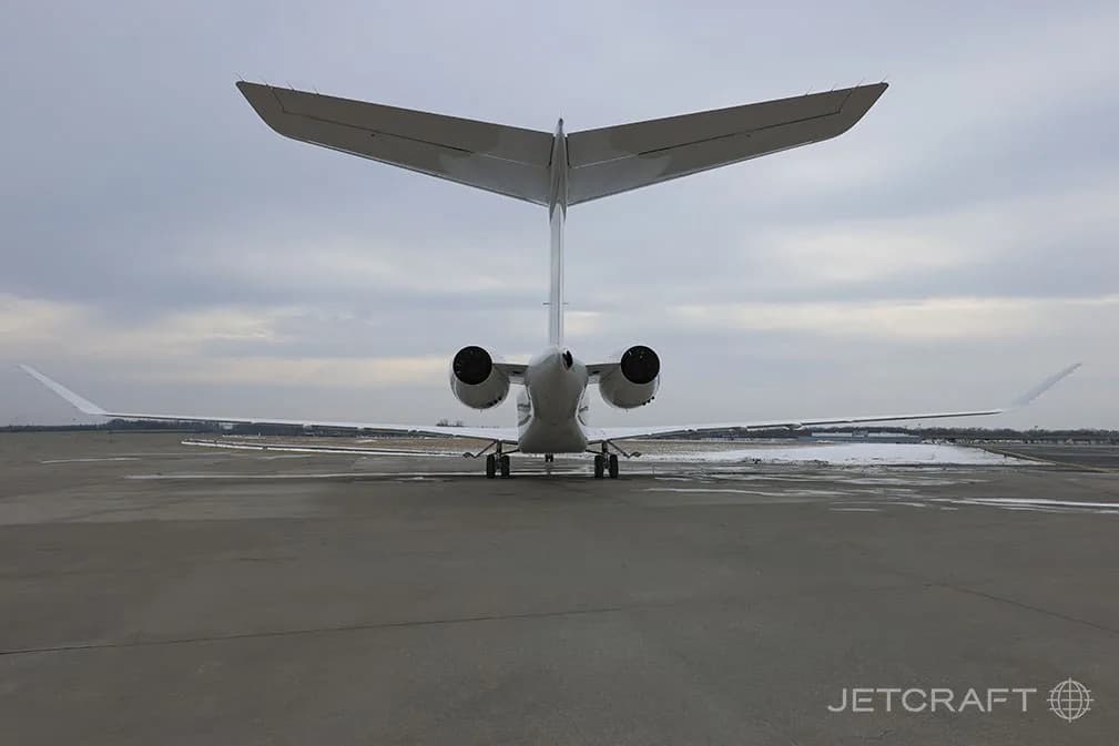 The rear view of a white Bombardier Global 7500 business jet parked on a tarmac with snow in the distance.