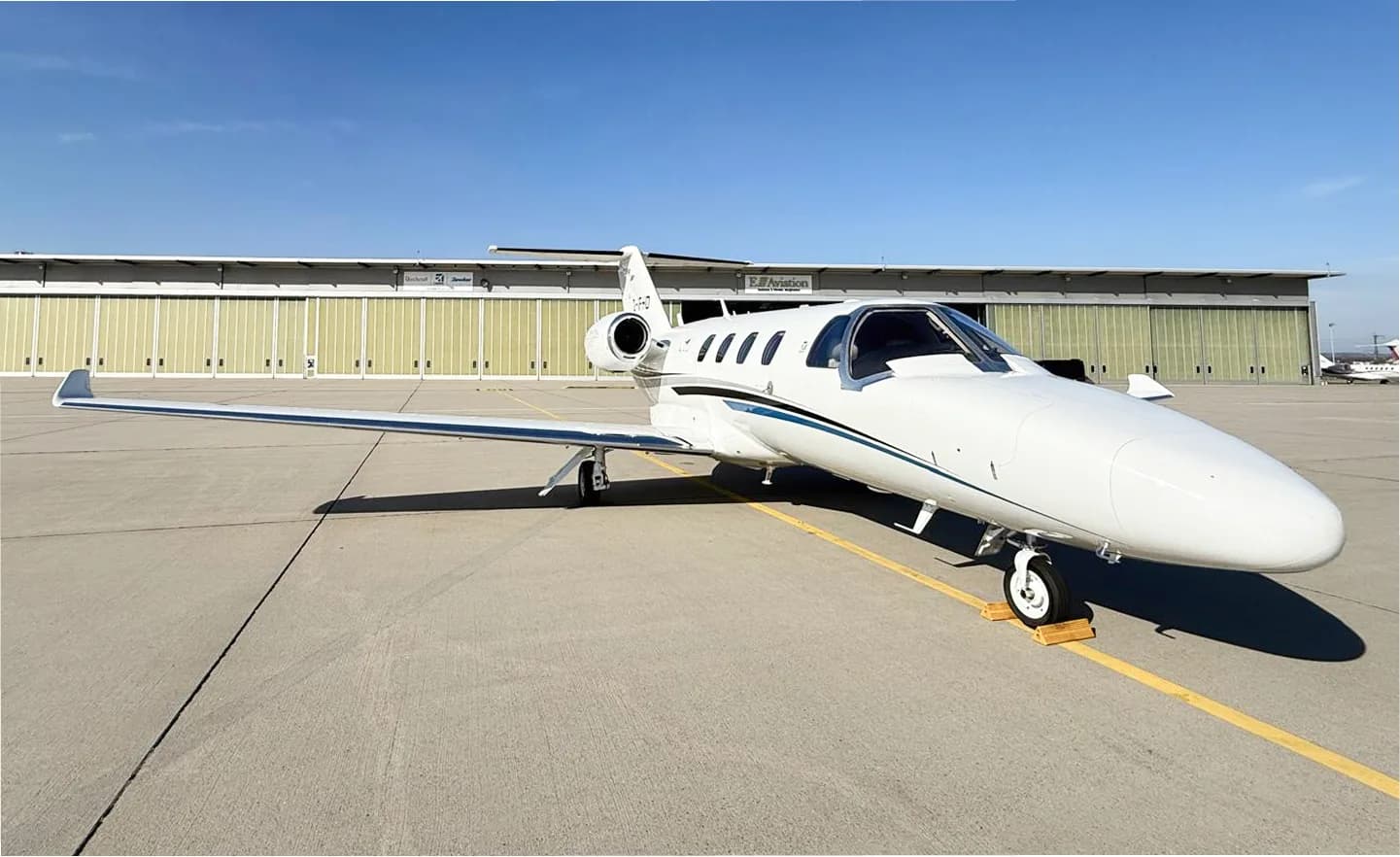 A white Cessna Citation business jet with a blue stripe is parked on an airport tarmac in front of hangars.