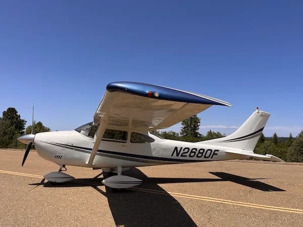 A white Cessna 182Q Skylane with a blue wing and grey stripes, parked on an asphalt surface under a clear blue sky.