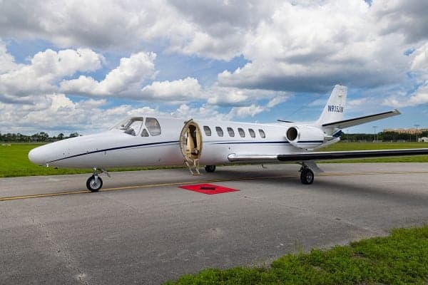 A Cessna Citation CJ3 aircraft with an open door is parked on the tarmac under a cloudy sky.
