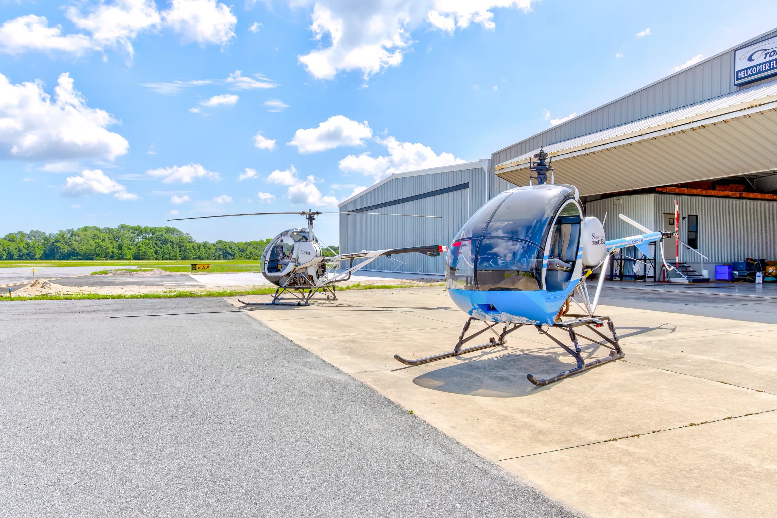 Two Schweizer 300CBi helicopters, N385TA (blue and white) and N124TA (black and white), parked on an airfield tarmac in front of hangars.