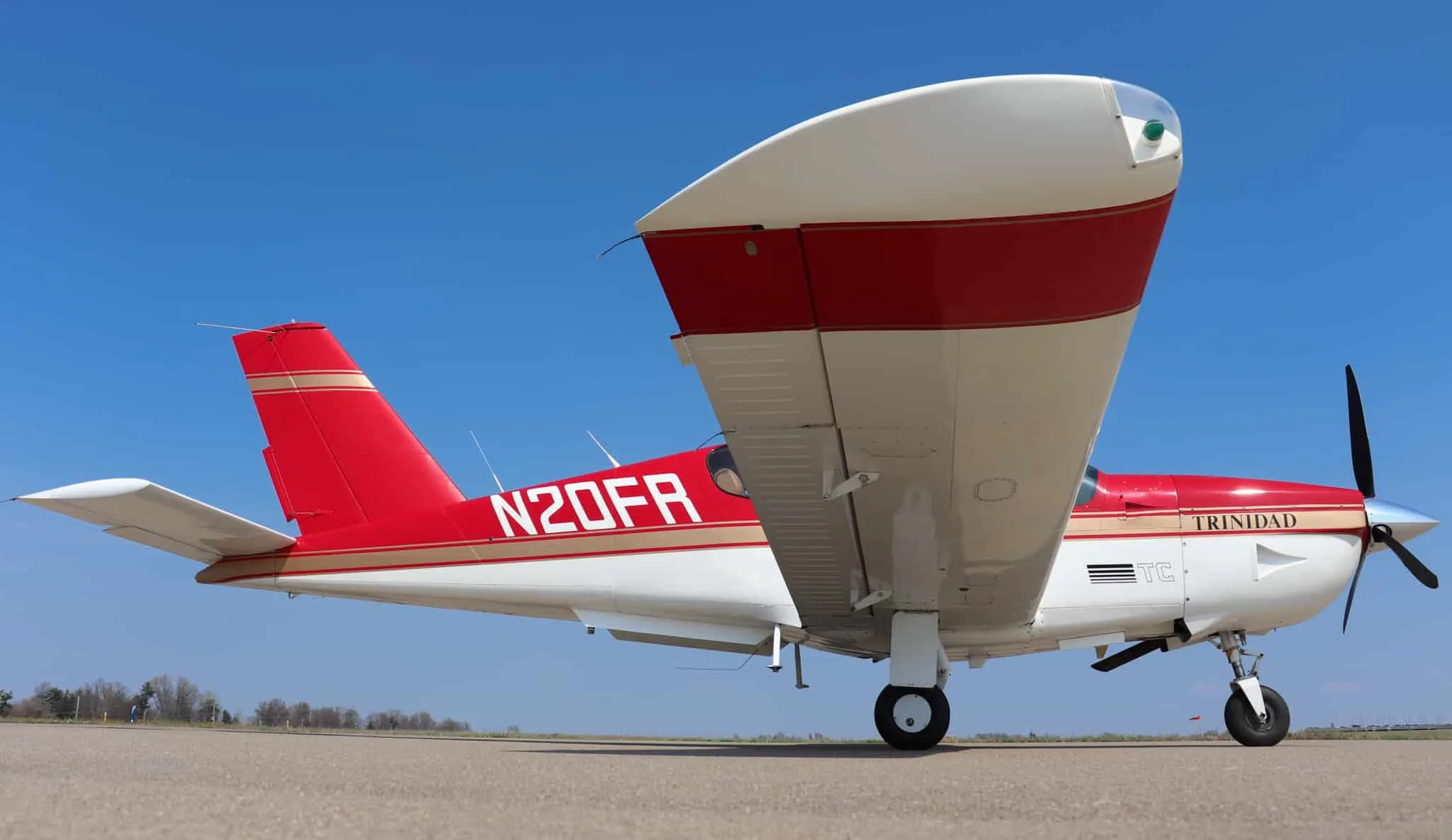A red and white Socata Trinidad aircraft with gold stripes is parked on an airport tarmac, viewed from the rear-right.