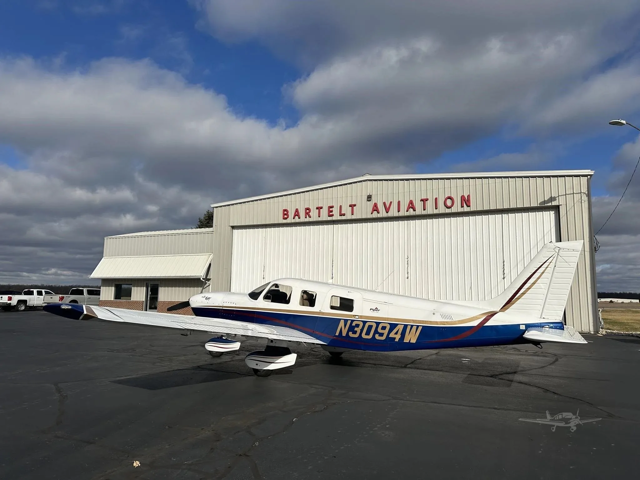 A three-quarter rear view of a blue and white Piper Malibu Mirage N3094W parked on an asphalt ramp in front of a Bartelt Aviation hangar.