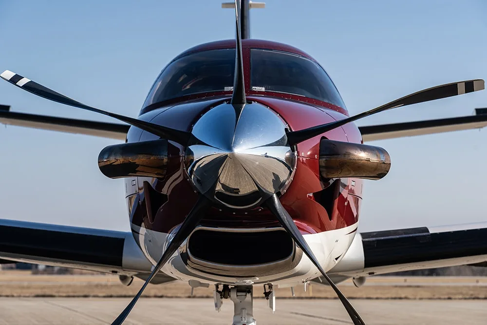 Head-on view of a maroon and white Daher TBM 900 aircraft, focusing on the propeller and nose.