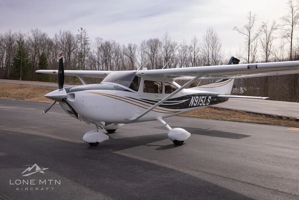 A three-quarter front view of a white and dark green Cessna Skylane N915LS with gold stripes, parked on an asphalt ramp.