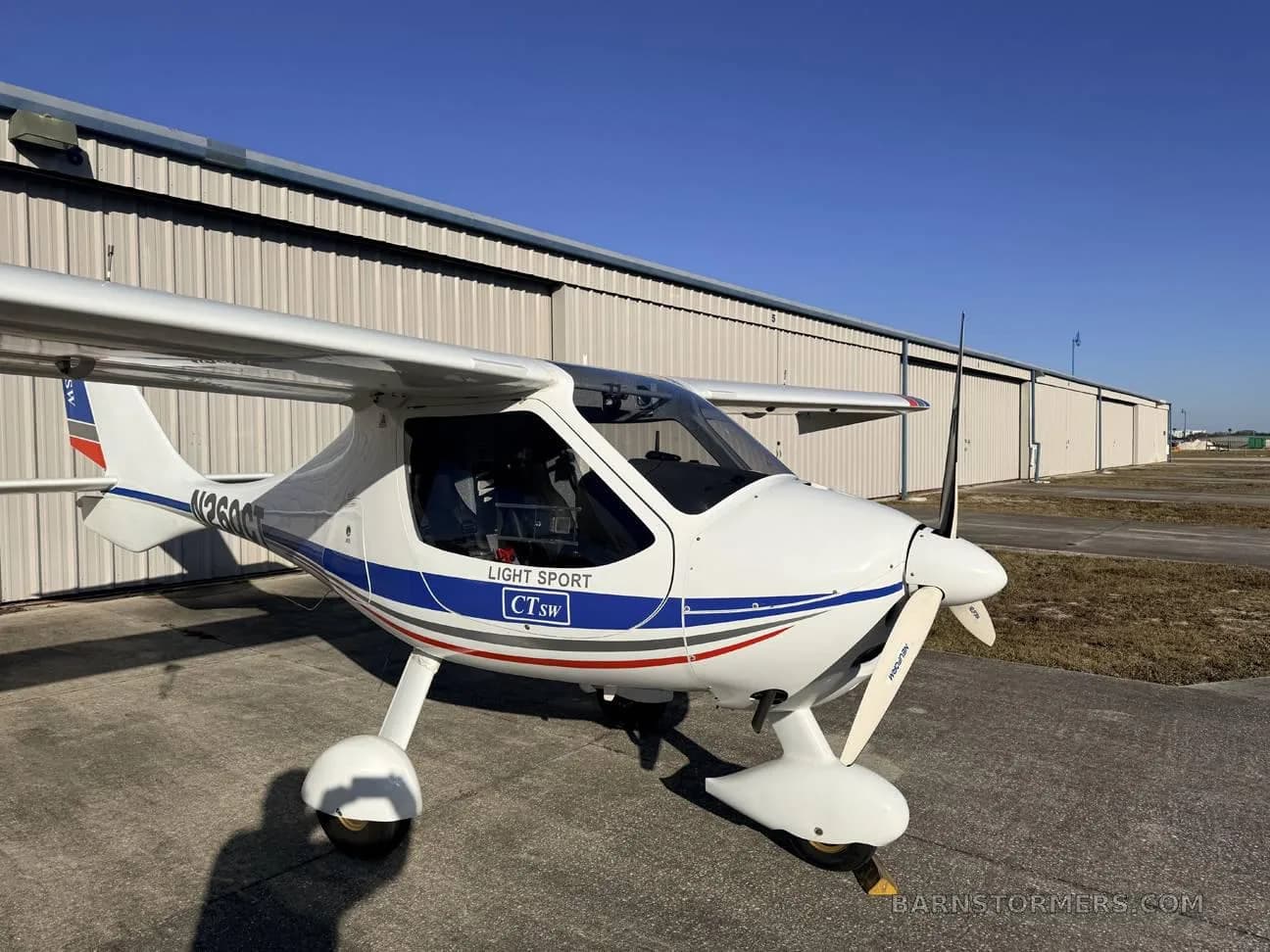 A white CTsw Light Sport aircraft with blue and red stripes, parked on a tarmac in front of a hangar under a clear blue sky.