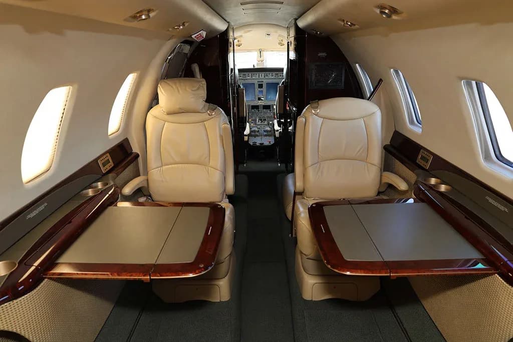 A view of the private jet cabin with beige leather seats and extended tables, looking towards the open cockpit door.