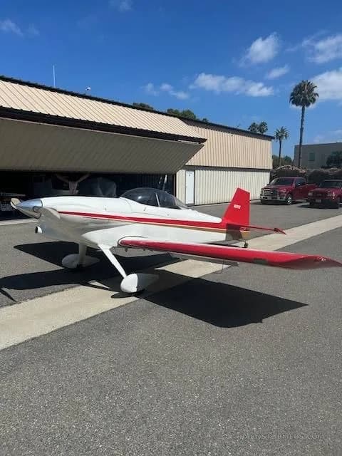 A white and red single-engine aircraft, possibly an experimental or aerobatic type, parked on an asphalt apron in front of a hangar.