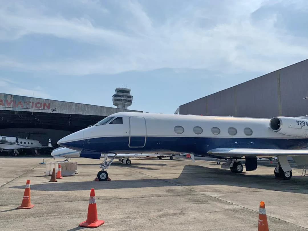 A Gulfstream G-IV business jet, registration N234LR, in a white and dark blue paint scheme, parked on a tarmac in front of a hangar and control tower.