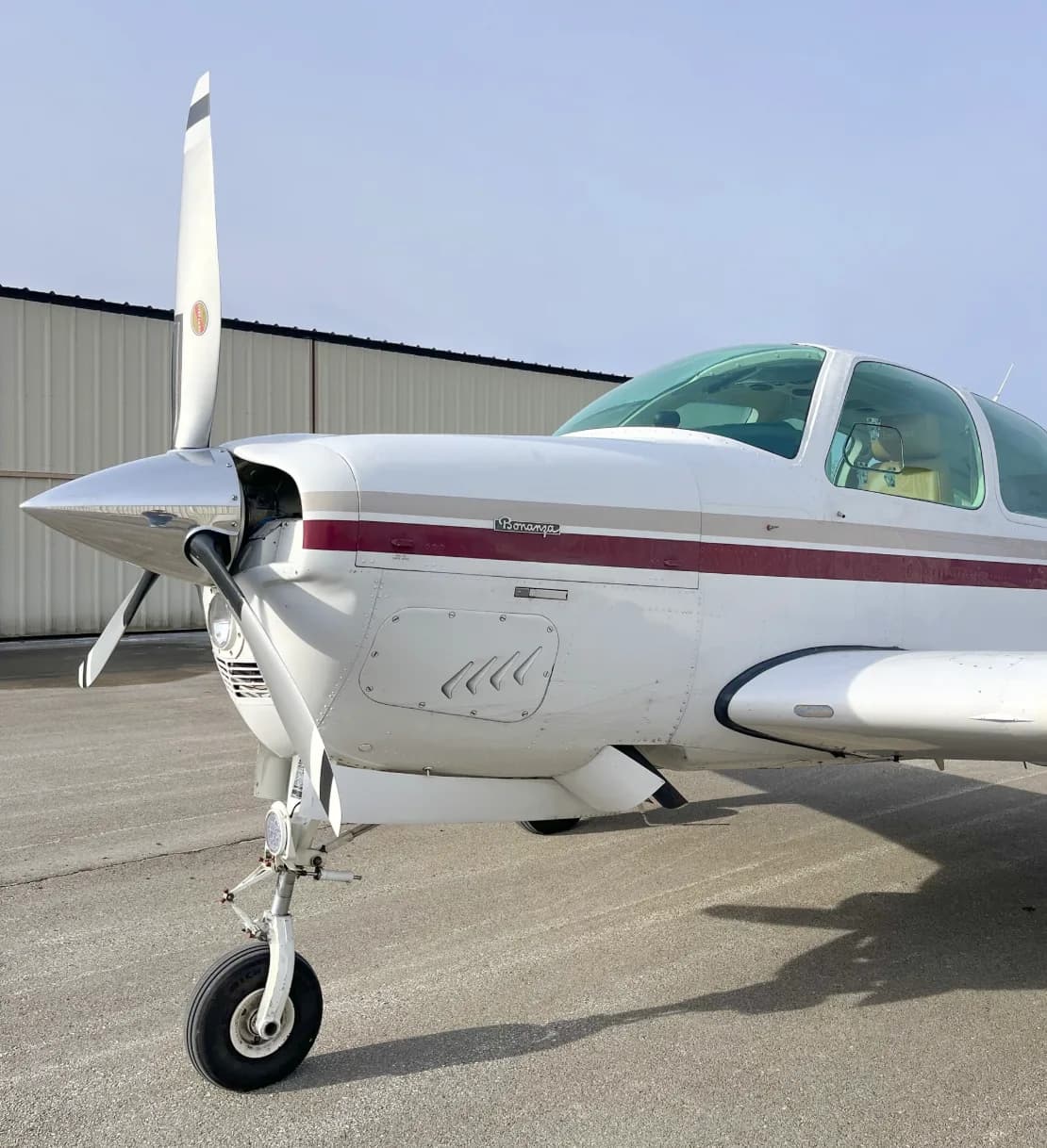 A close-up side view of the nose and propeller of a white and maroon Beechcraft Bonanza aircraft.