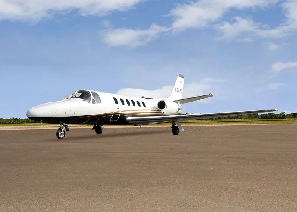 A white and black Cessna Citation business jet N941RJ with gold stripes is parked on a tarmac under a partly cloudy sky, viewed from the front-left.