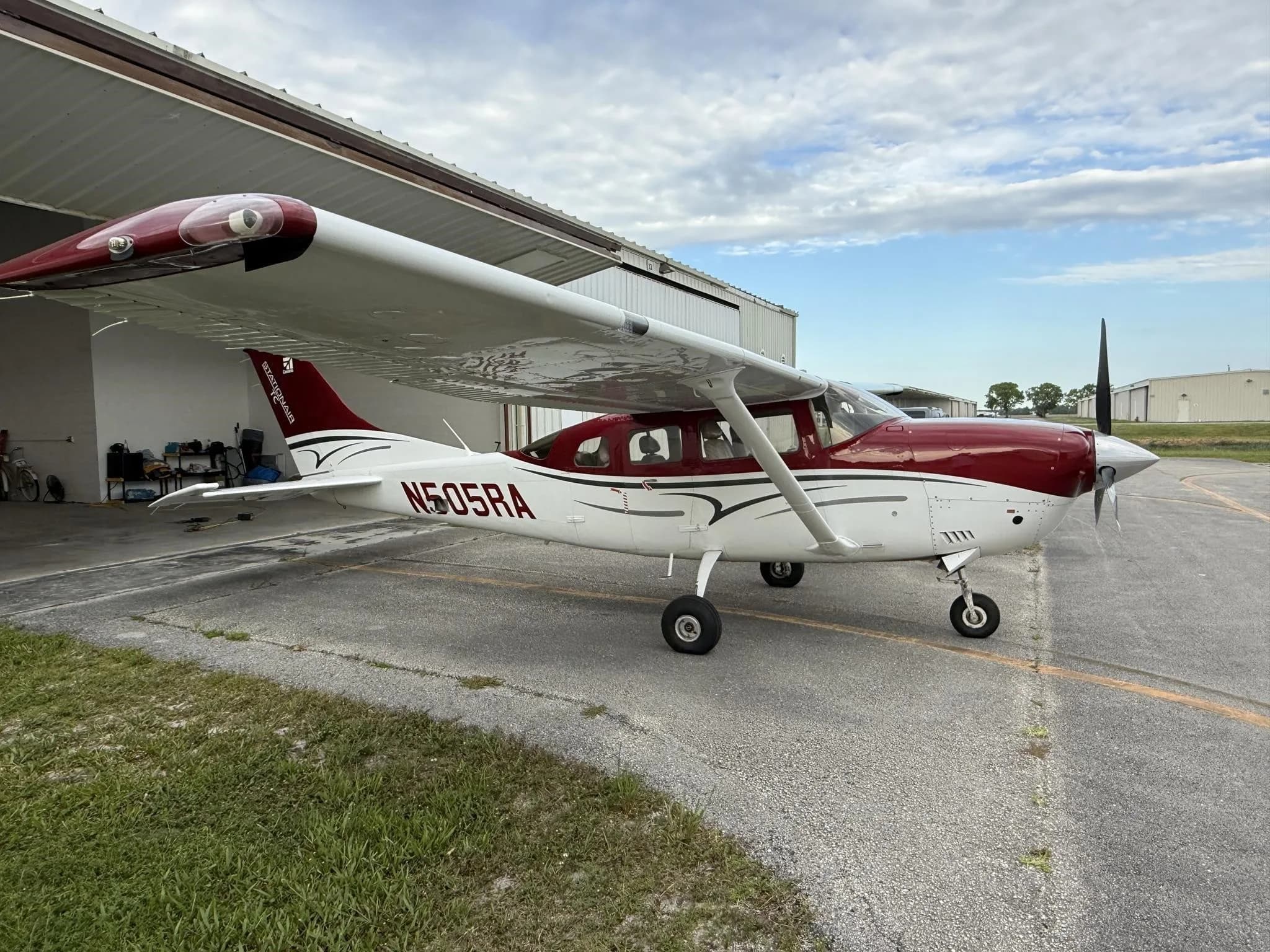 A white and red Cessna 182 aircraft, registration N505RA, parked on an asphalt ramp next to a hangar under a cloudy sky.