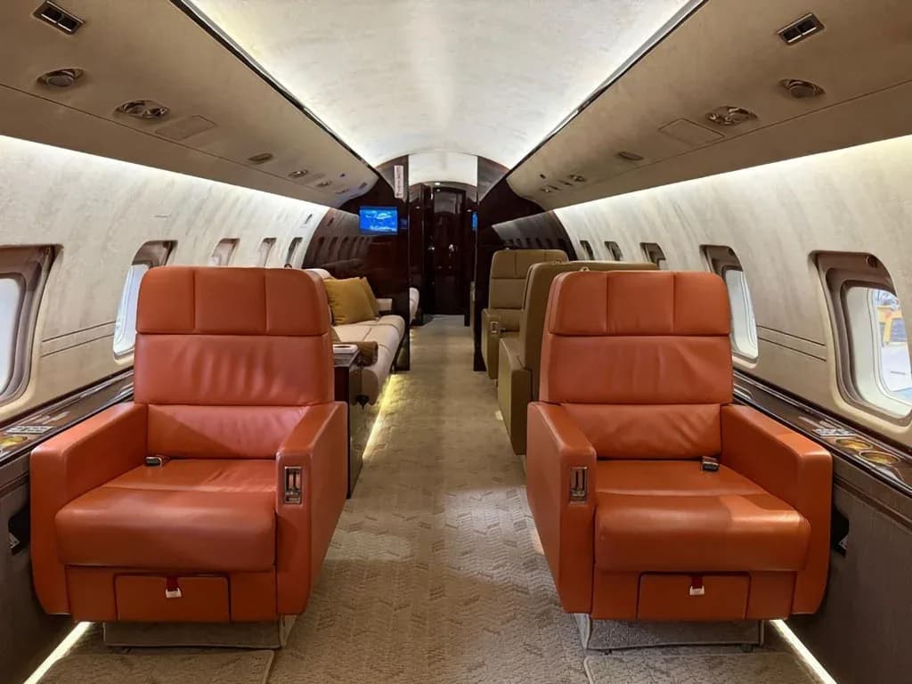 The luxurious interior cabin of a private jet features orange leather seats and a light-colored carpet, looking towards the rear.