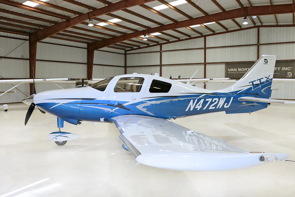 A white and blue Cessna TTx aircraft, registration N472WJ, is parked in a hangar, viewed from the side.