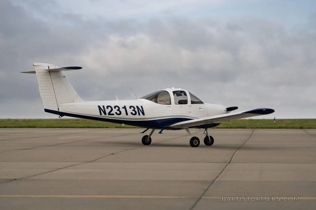 A white and blue single-engine propeller aircraft with registration N2313N is parked on an asphalt runway under a cloudy sky, viewed from the side.