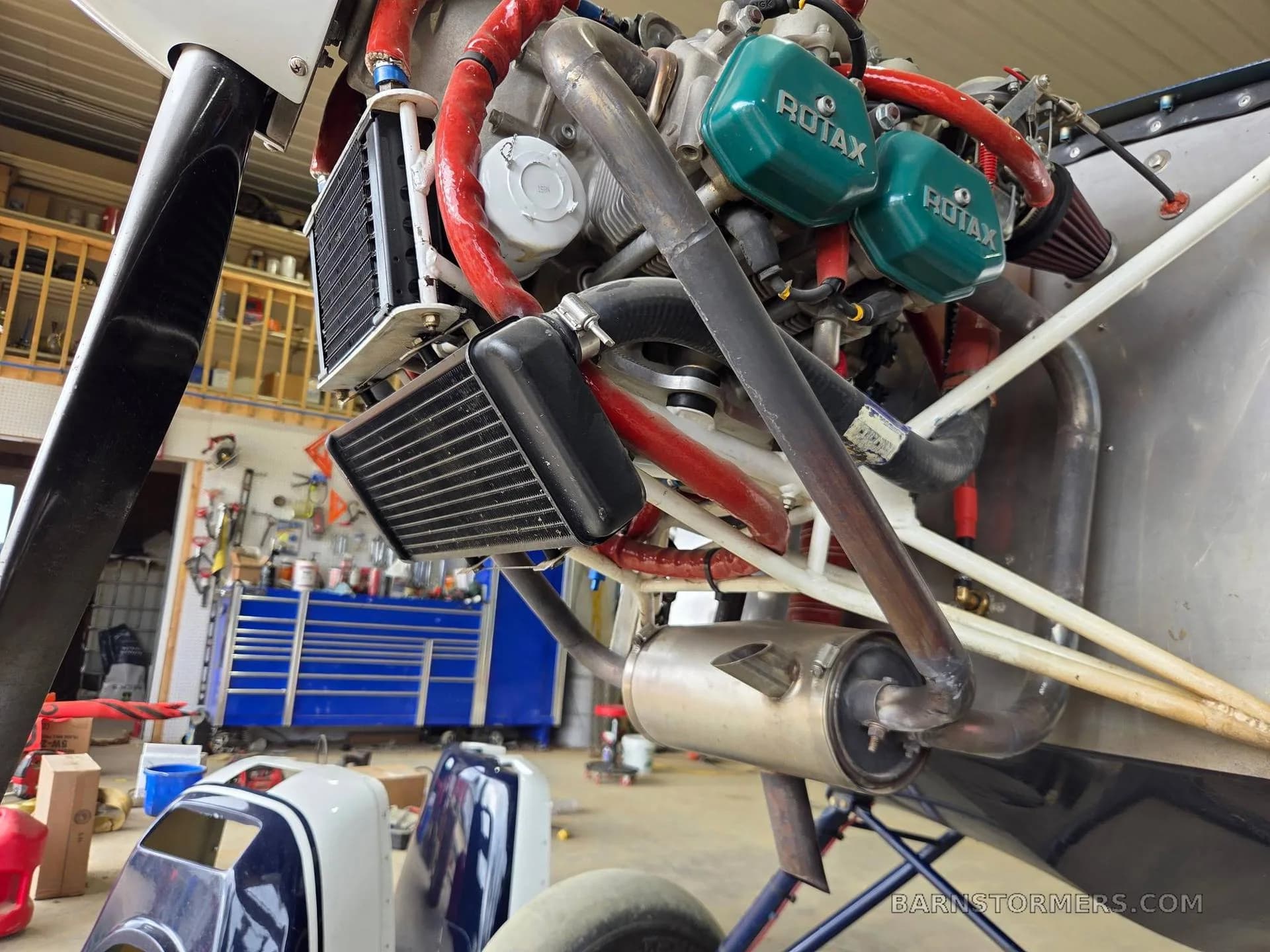 A detailed view of the Rotax aircraft engine, showing the exhaust, cooling fins, and propeller in a hangar.