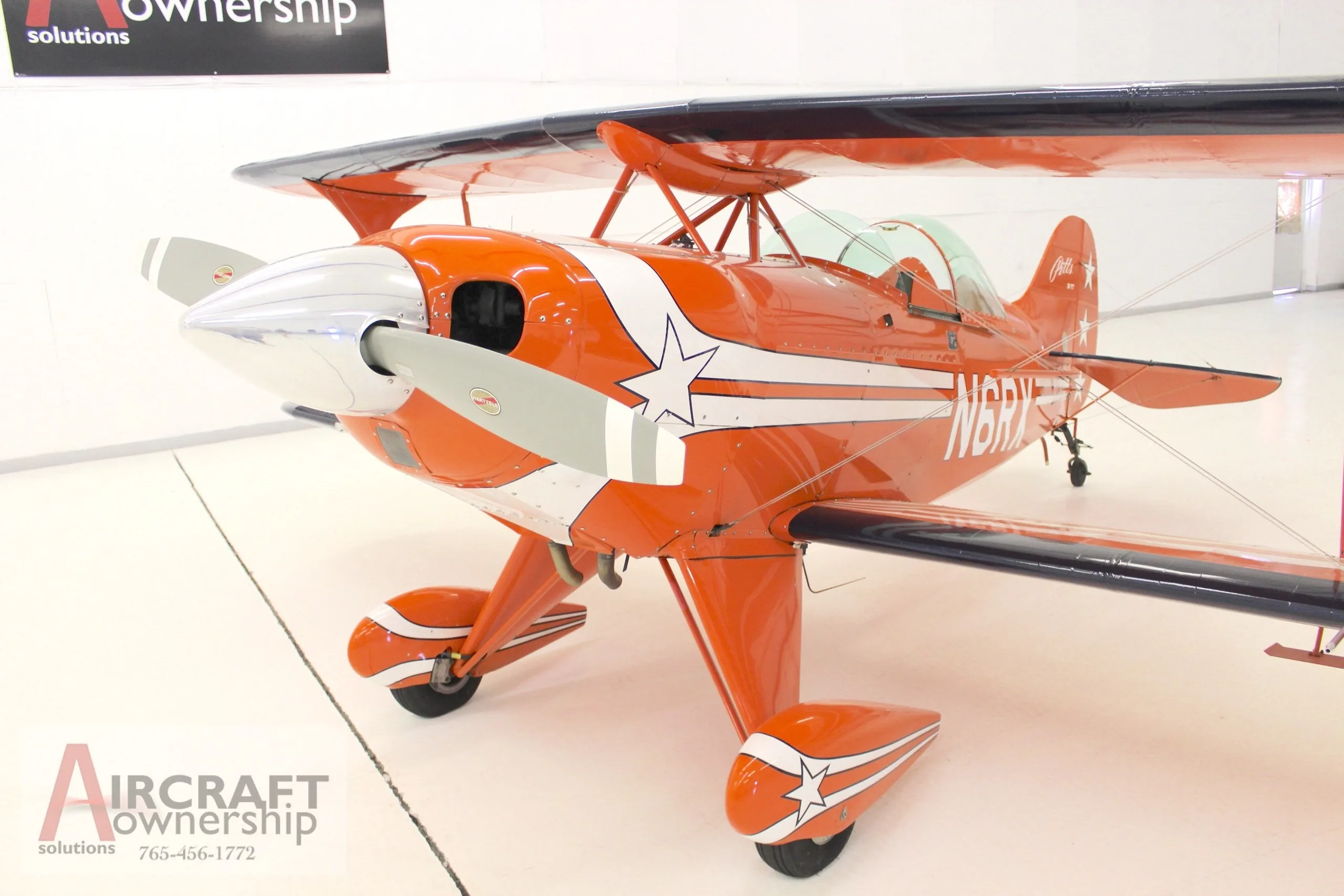 A front-quarter view of an orange Pitts S-1T biplane with white stripes and stars, showing the propeller and landing gear.