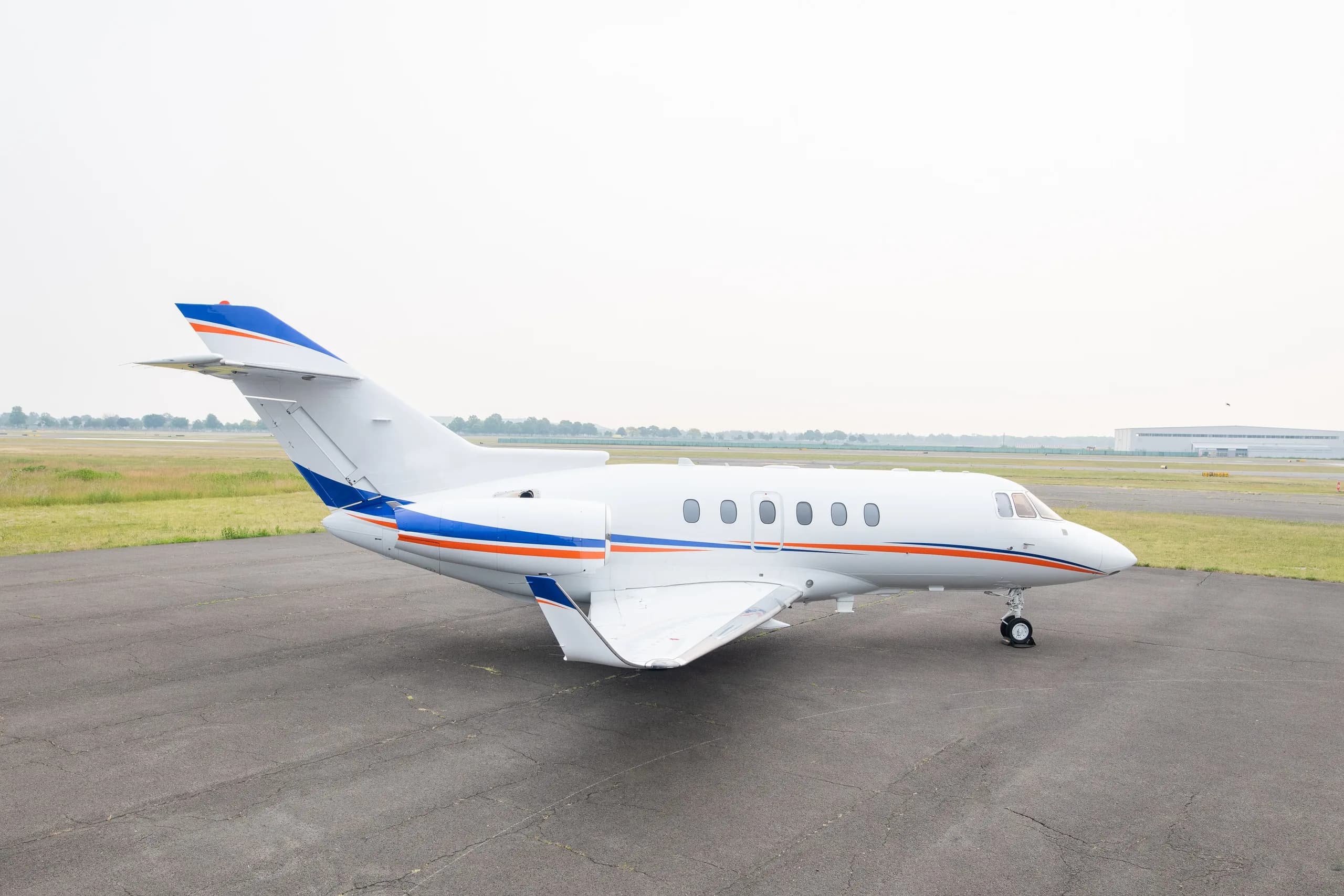 A white business jet with blue and orange stripes on the tarmac, viewed from the rear-left.