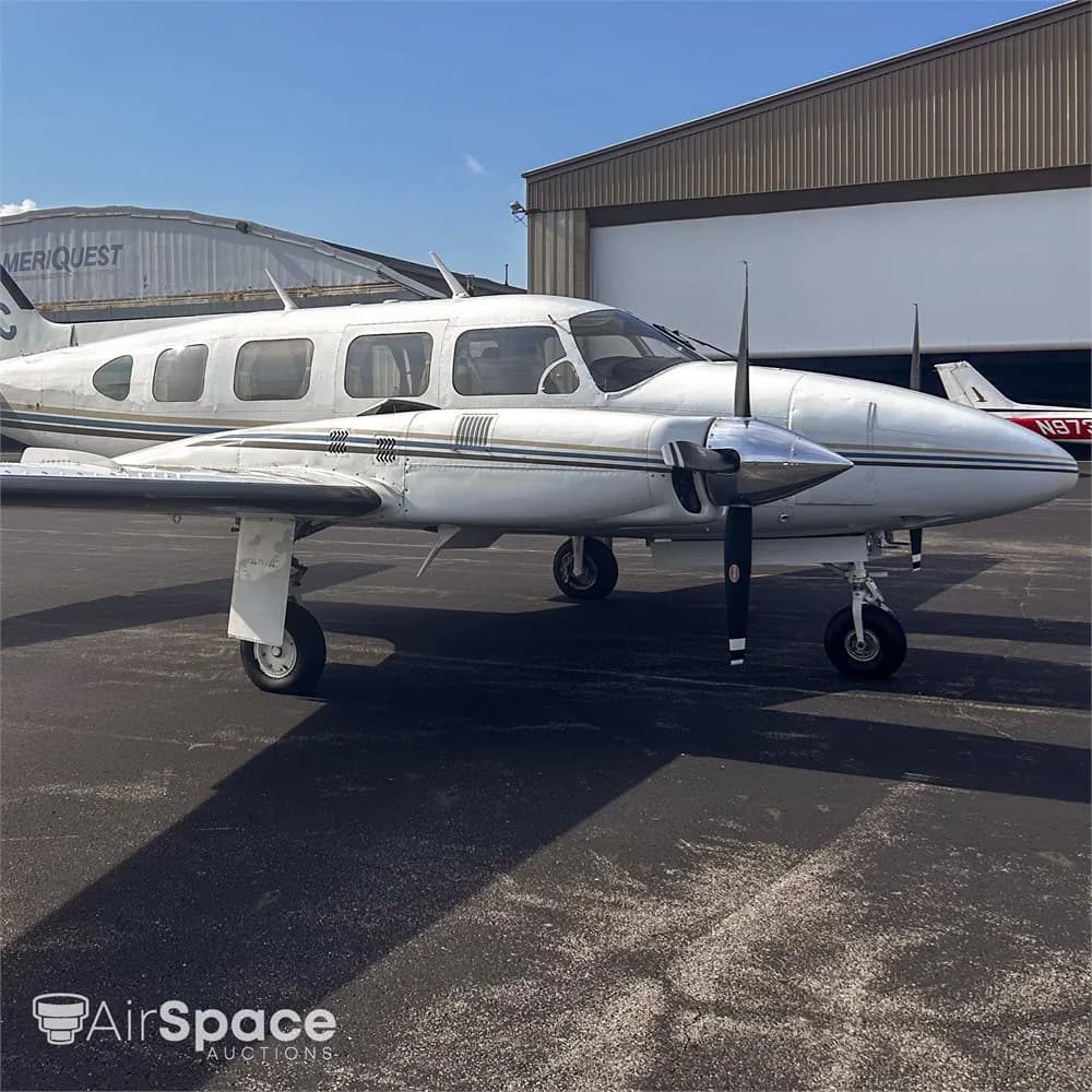 A white Piper Navajo aircraft with blue and gold stripes parked on an asphalt tarmac, viewed from the front-right.