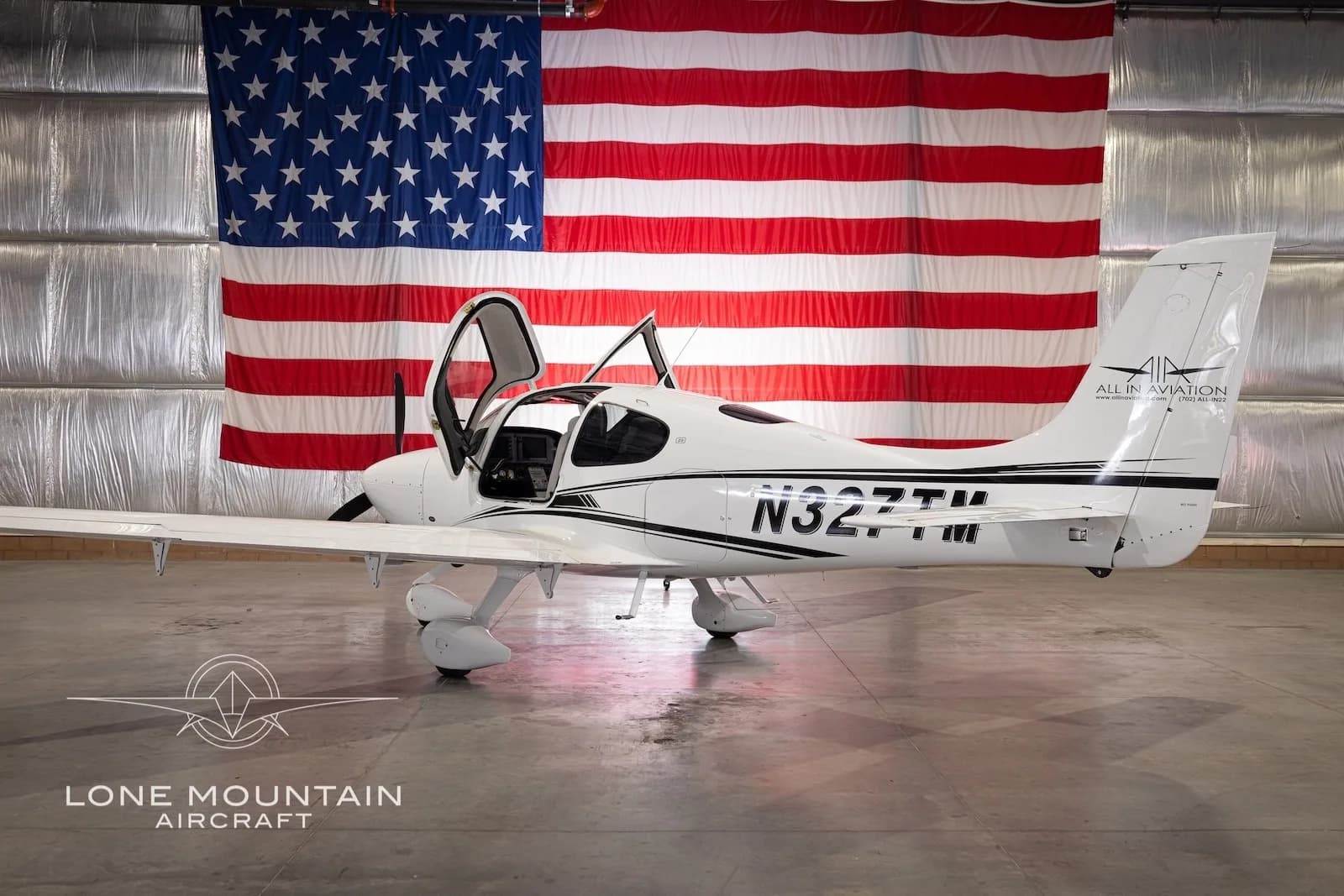 A white Cirrus SR22 aircraft with black stripes and registration N327TM, parked in a hangar with an American flag backdrop, with its gull-wing doors open.