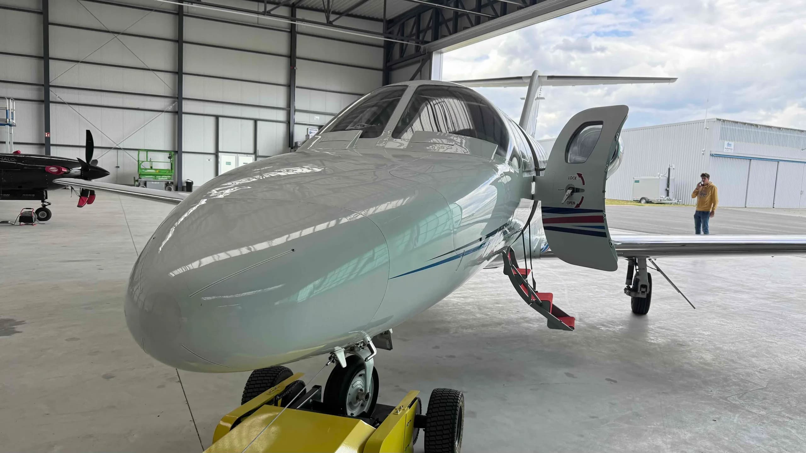 A white private jet, likely a HondaJet, with its main door open and red steps extended, parked inside a hangar with another aircraft partially visible.