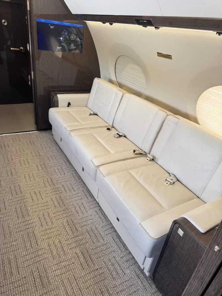 A close-up of a long, light-colored leather divan in the Gulfstream cabin, with a screen displaying a map on the wall.