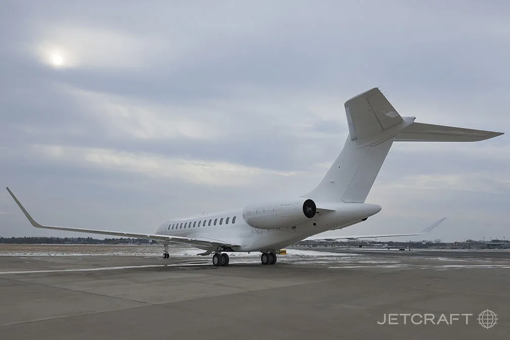 A white Bombardier Global 7500 business jet is seen from the rear-left, parked on a snowy tarmac under a cloudy sky.