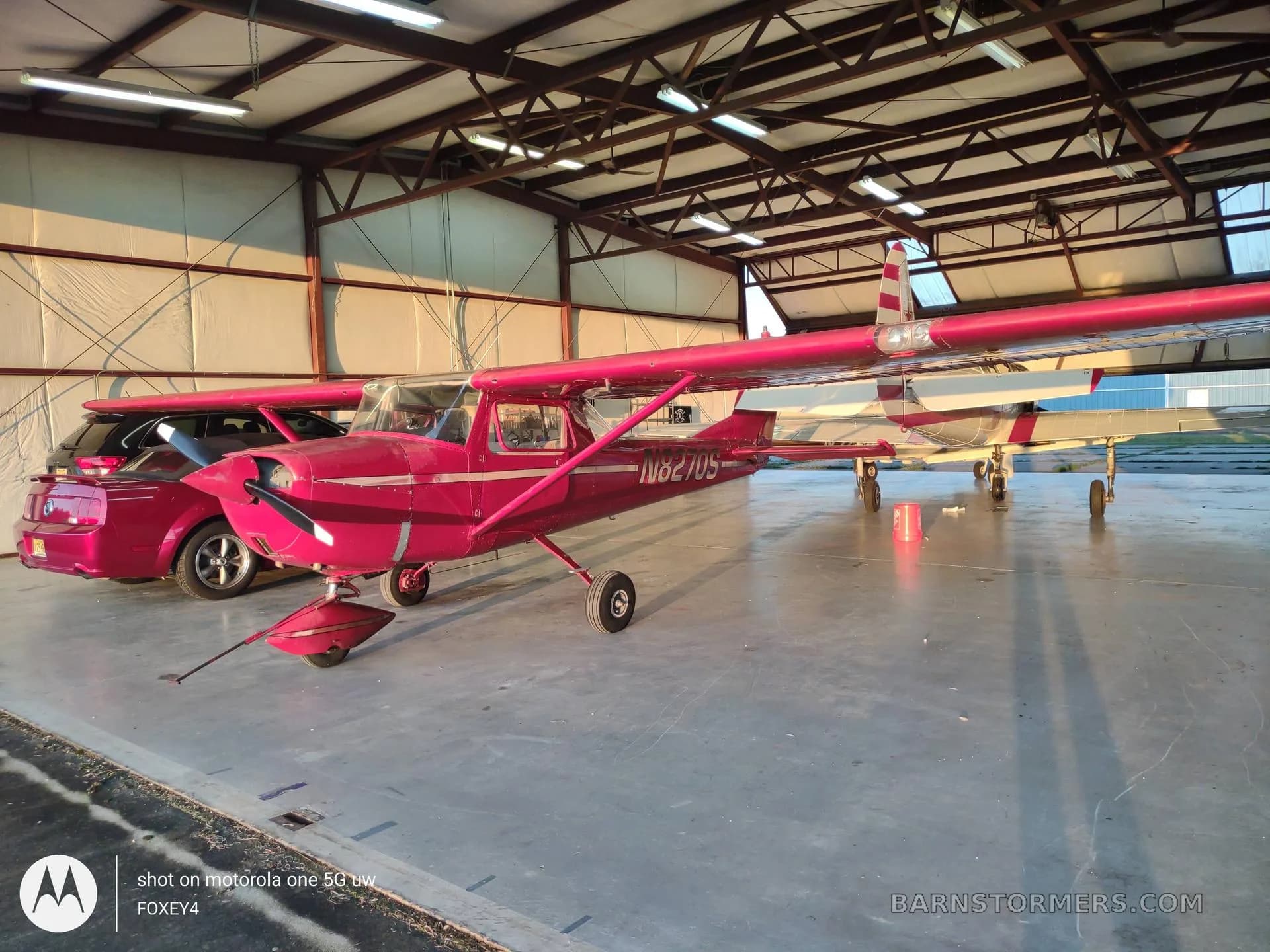 A red Cessna 150 or similar light aircraft with registration N8270S is parked inside a hangar next to a red car.