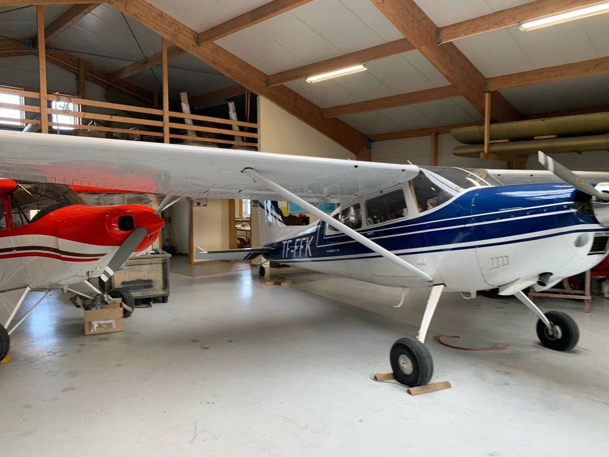 A white and blue Cessna 180 aircraft, registration TF-FFK, parked next to a red aircraft inside a hangar.