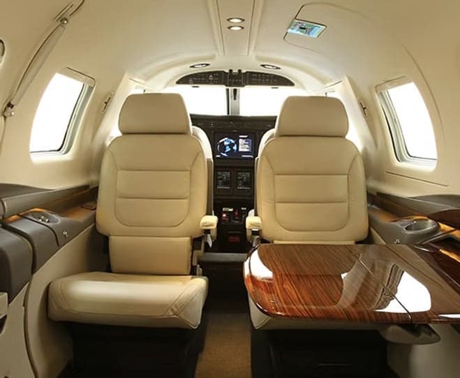 The luxurious beige leather interior of an aircraft cabin with two forward-facing seats and a wooden table.