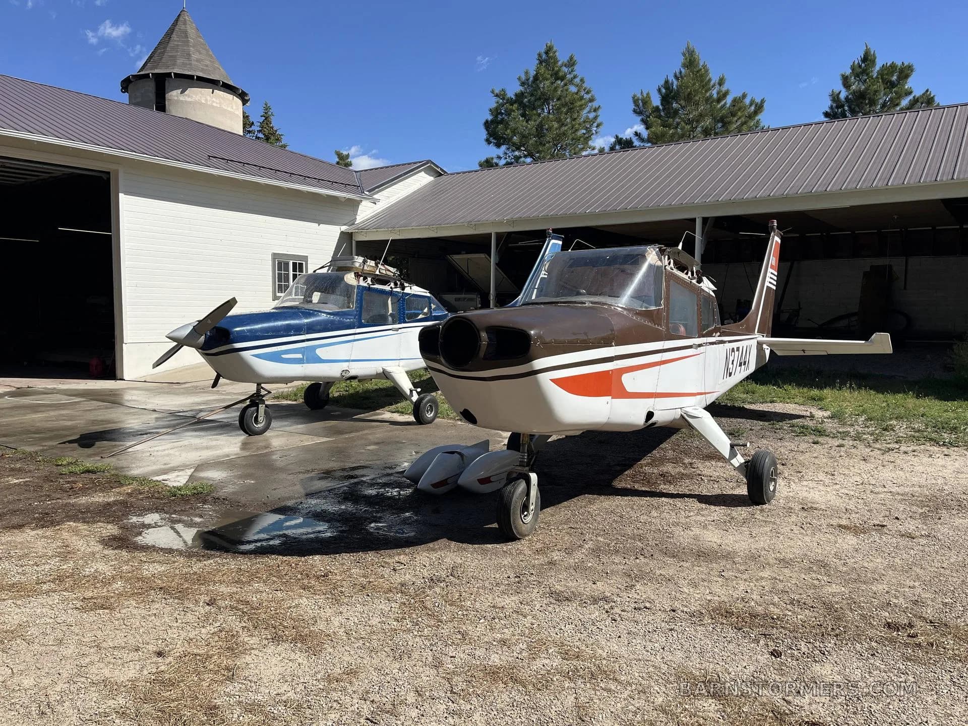 Two single-engine propeller aircraft, one blue and white and one brown and white, parked on a dirt and concrete area in front of a hangar.