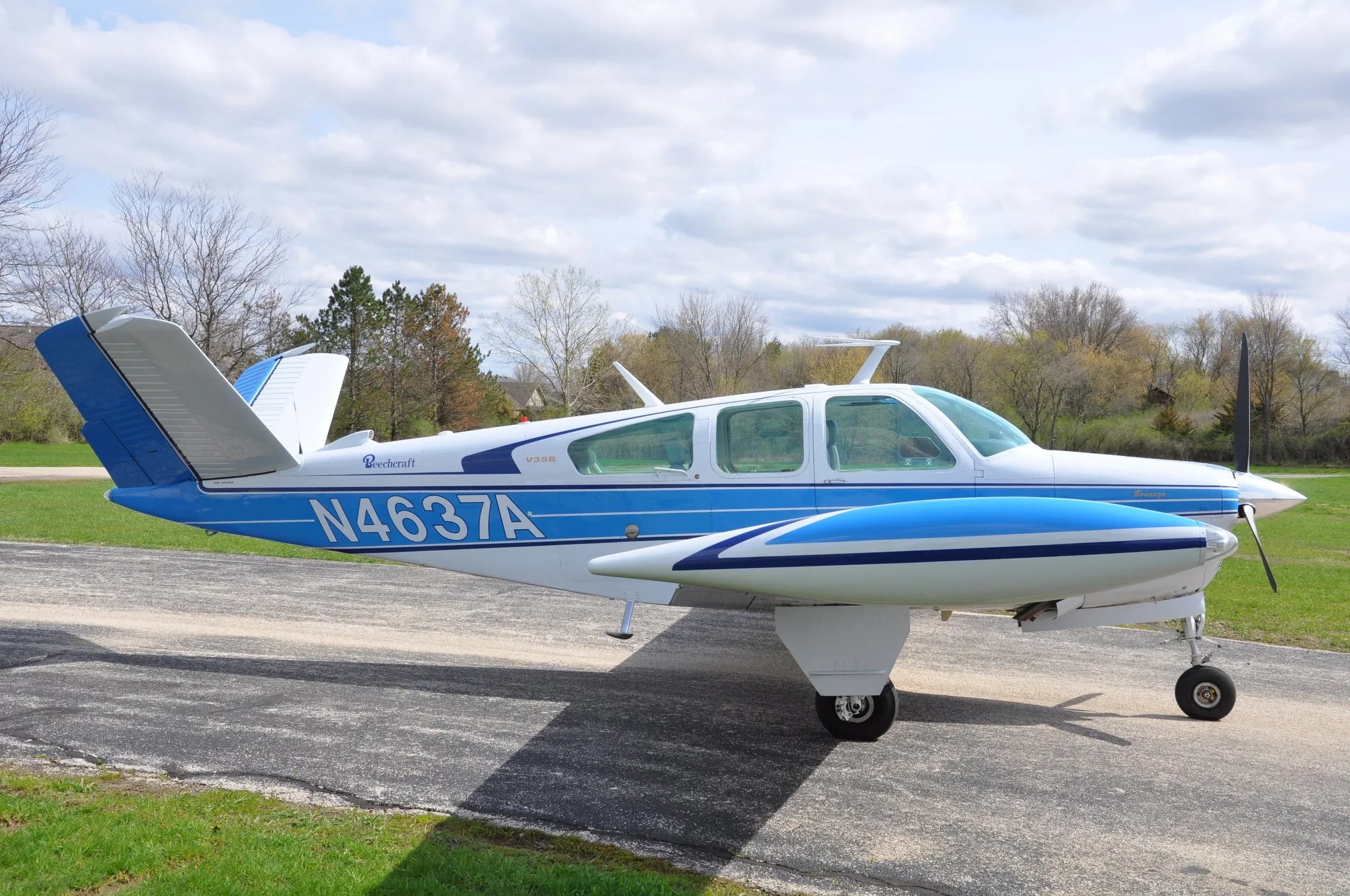 A white and blue Beechcraft Bonanza with registration N4637A, viewed from the side, parked on an asphalt runway under a partly cloudy sky.