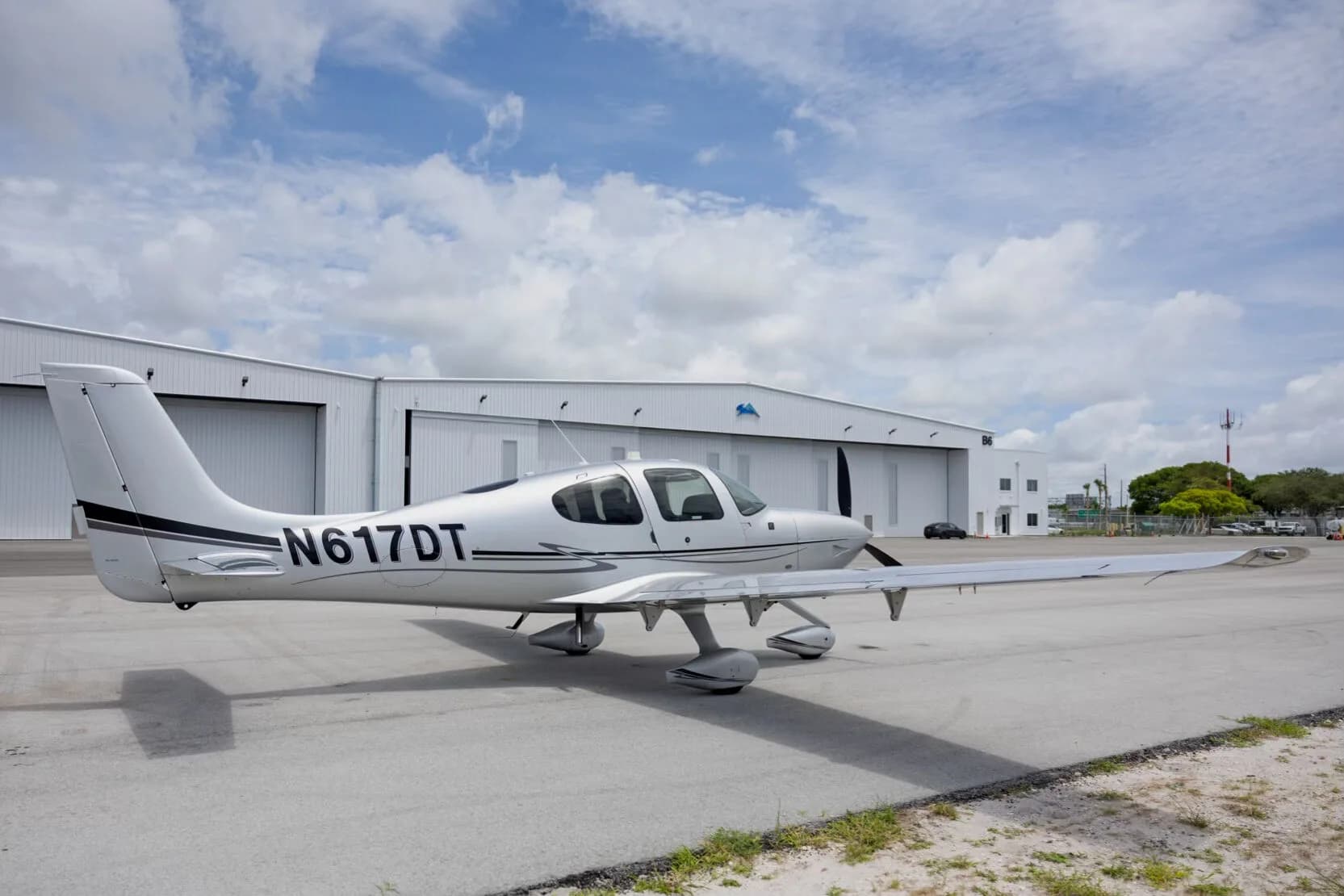 A side view of the silver Cirrus SR22 aircraft with tail number N617DT parked on an tarmac in front of hangars.