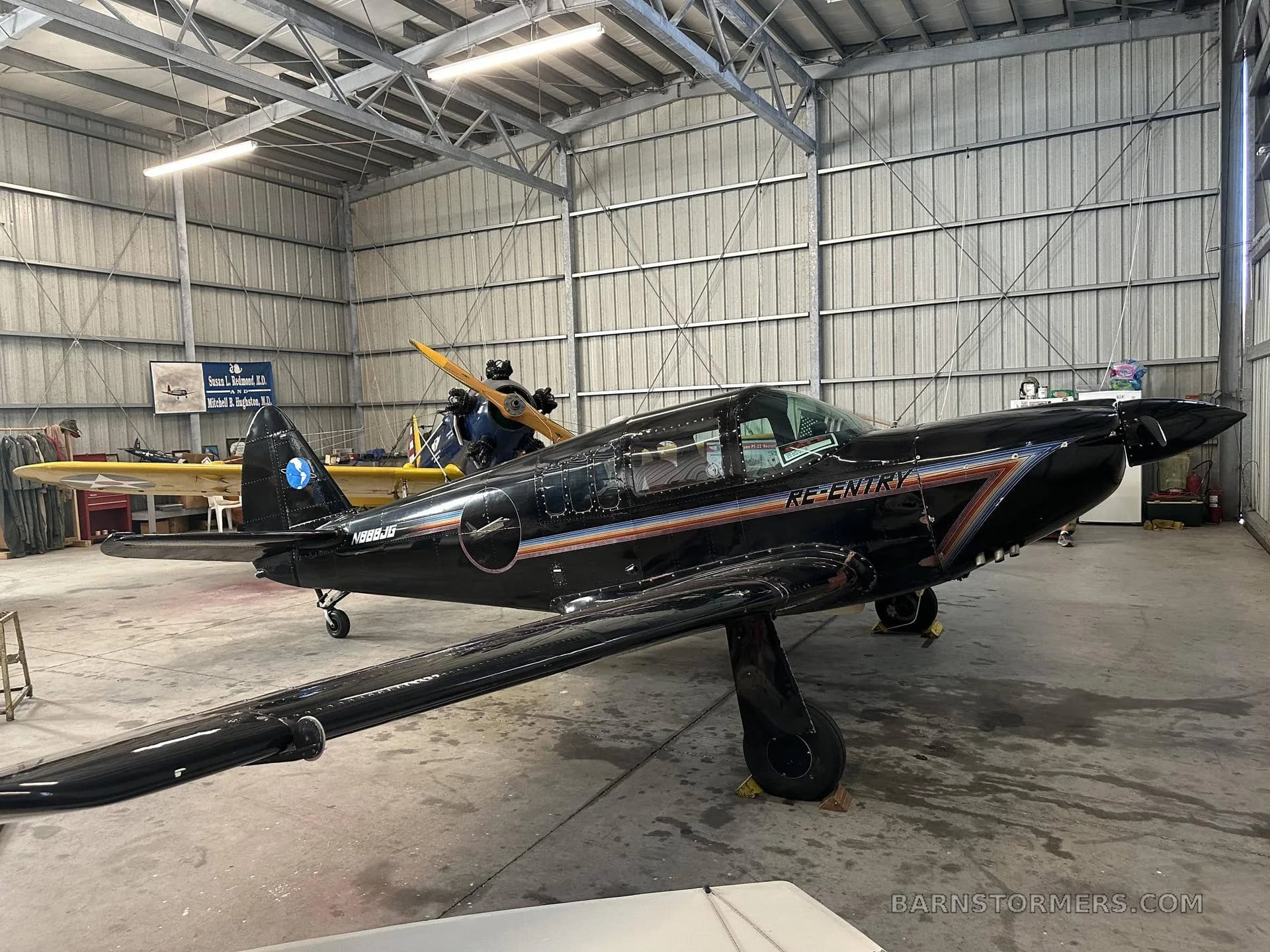 A black single-engine aircraft with 'RE-ENTRY' and registration N888JG on its side, parked inside a hangar with another yellow aircraft in the background.