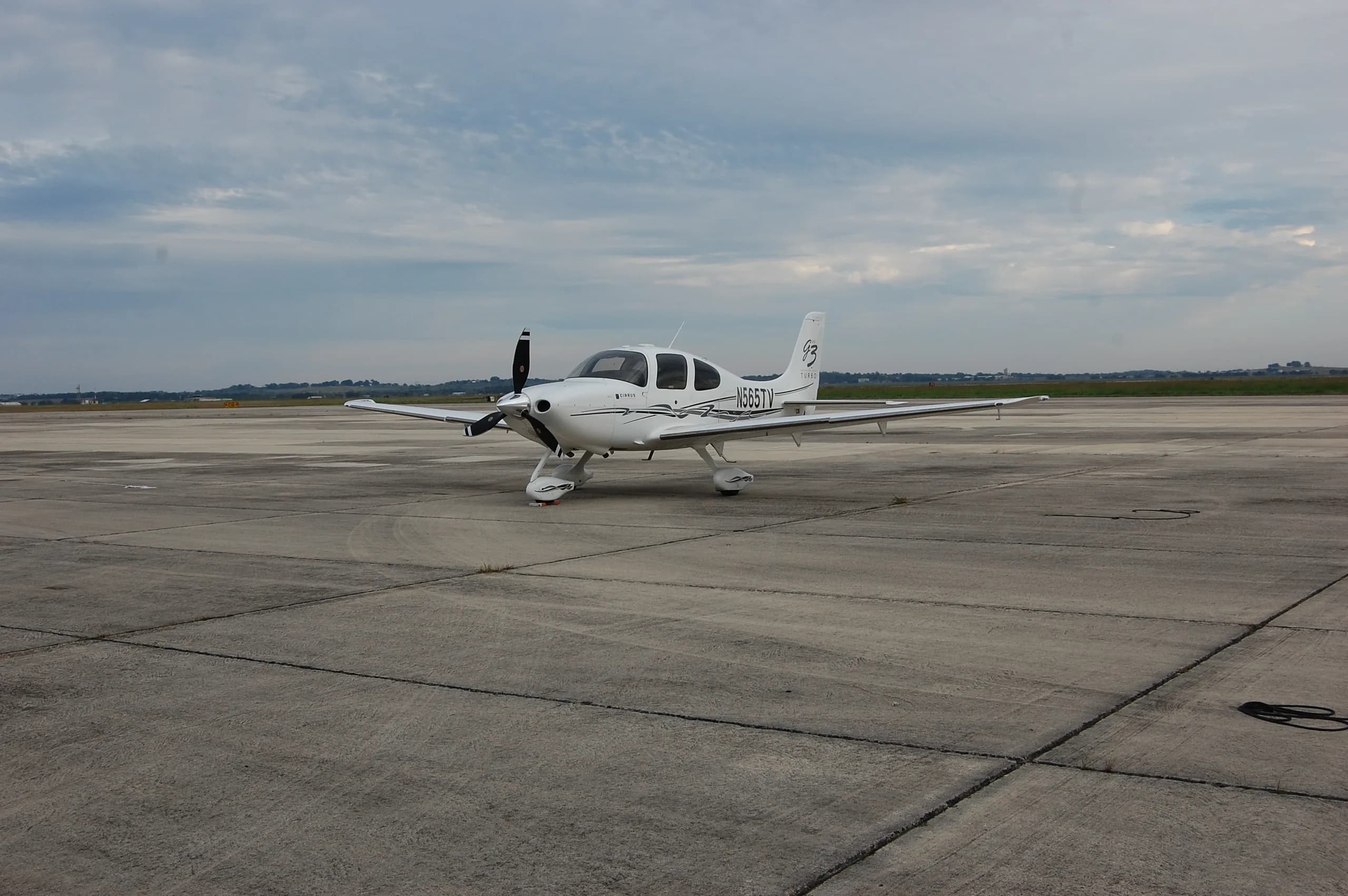 A white Cirrus SR22T aircraft, N565TV, parked on a concrete tarmac under a cloudy sky.