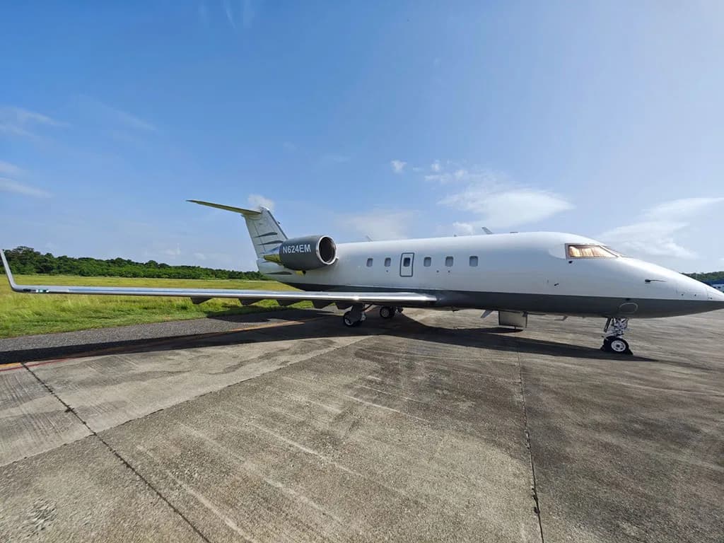 A side view of the white and grey Bombardier Challenger 604 business jet, N624EM, parked on an airfield under a blue sky.