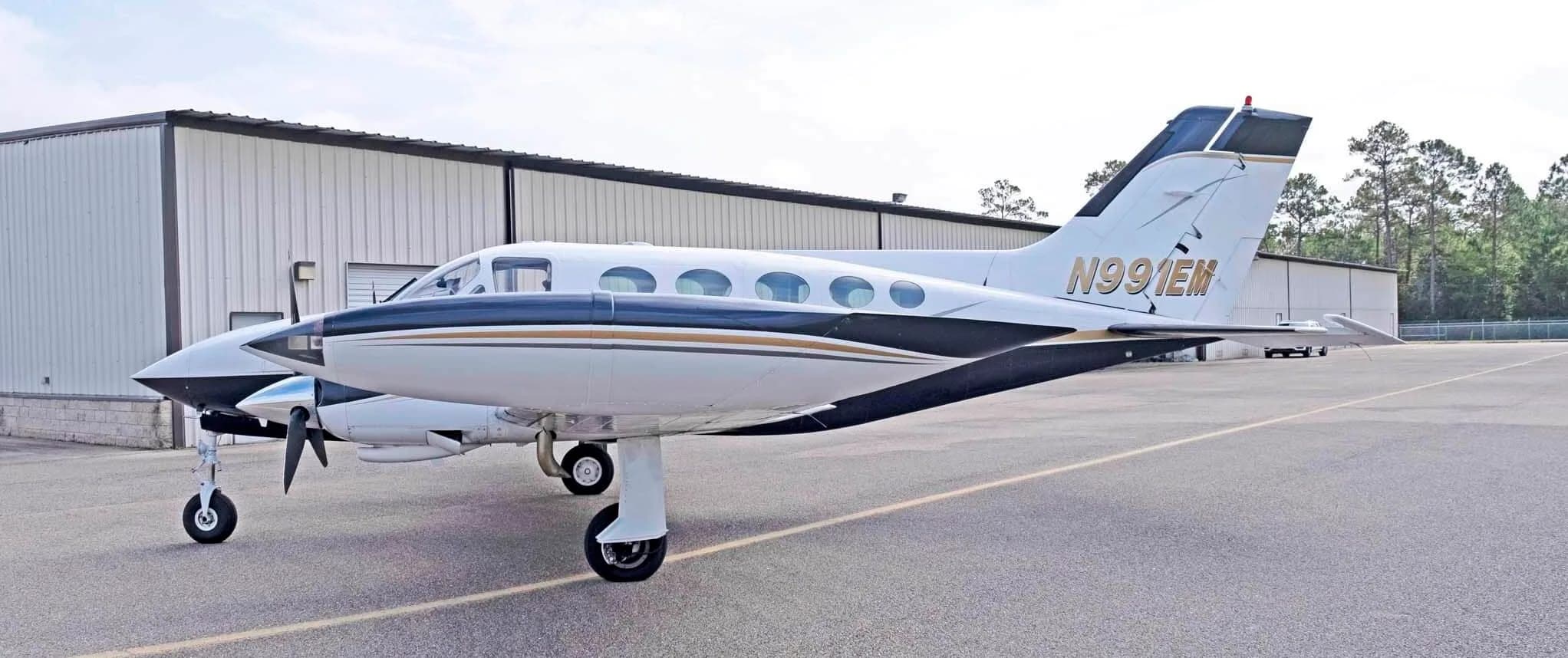 A side view of the white and dark blue Cessna 414 Chancellor, registration N991EM, parked in front of a hangar.