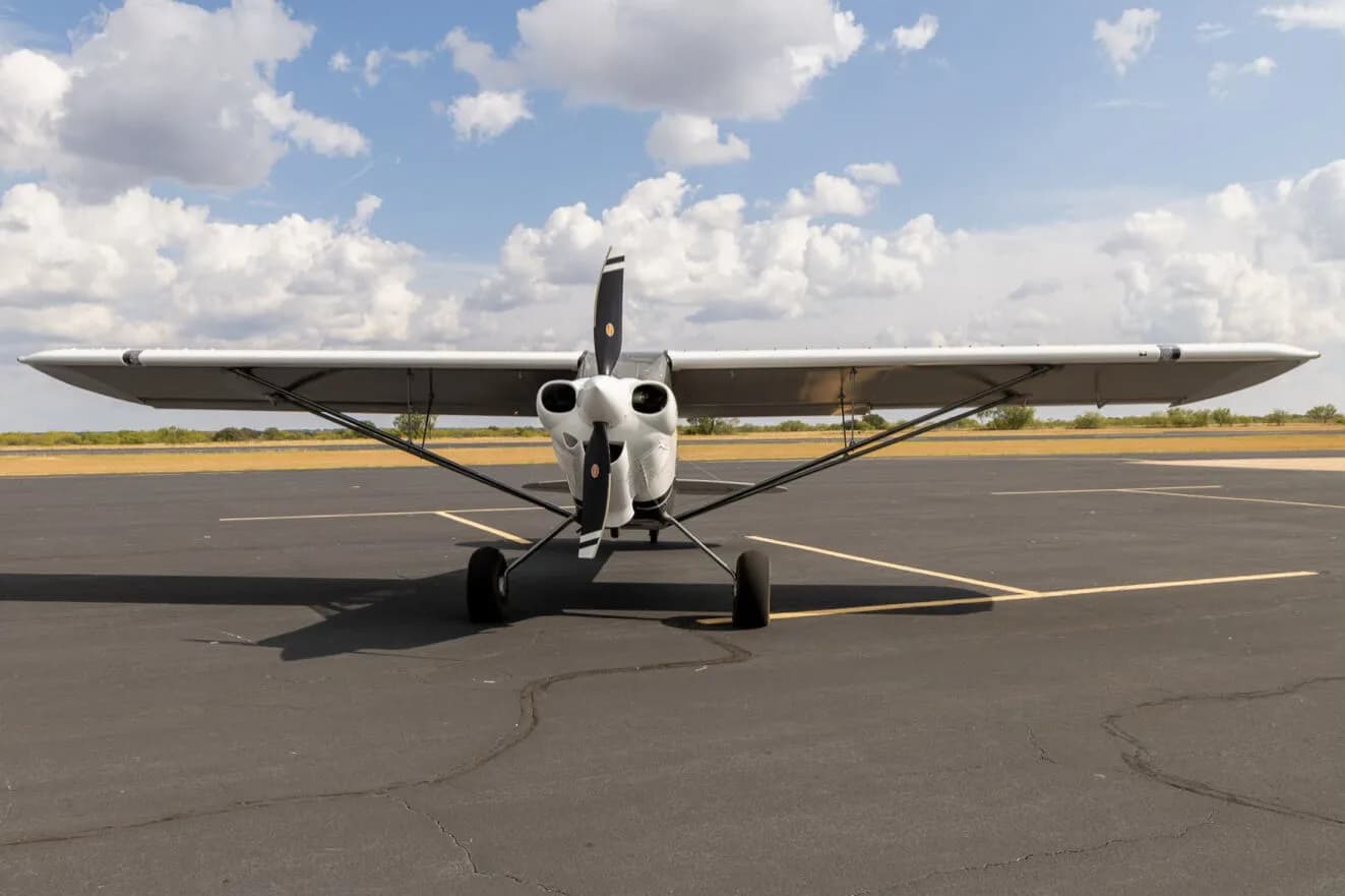 A head-on view of the white X-Cub aircraft with a black propeller, parked on an asphalt runway under a blue sky with clouds.