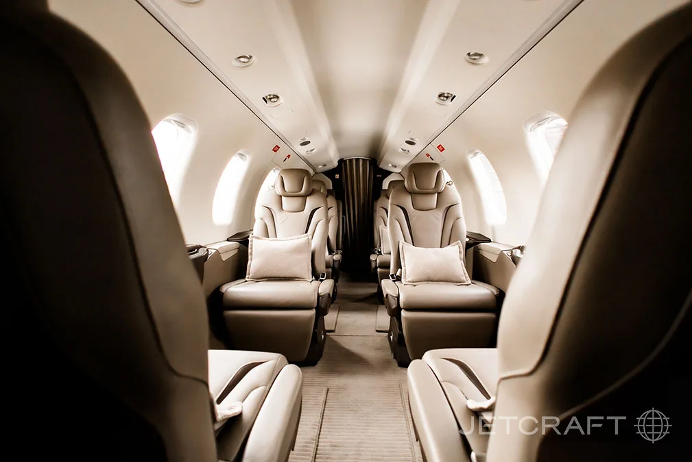 A view down the aircraft cabin aisle shows light brown leather seats with small pillows on either side.