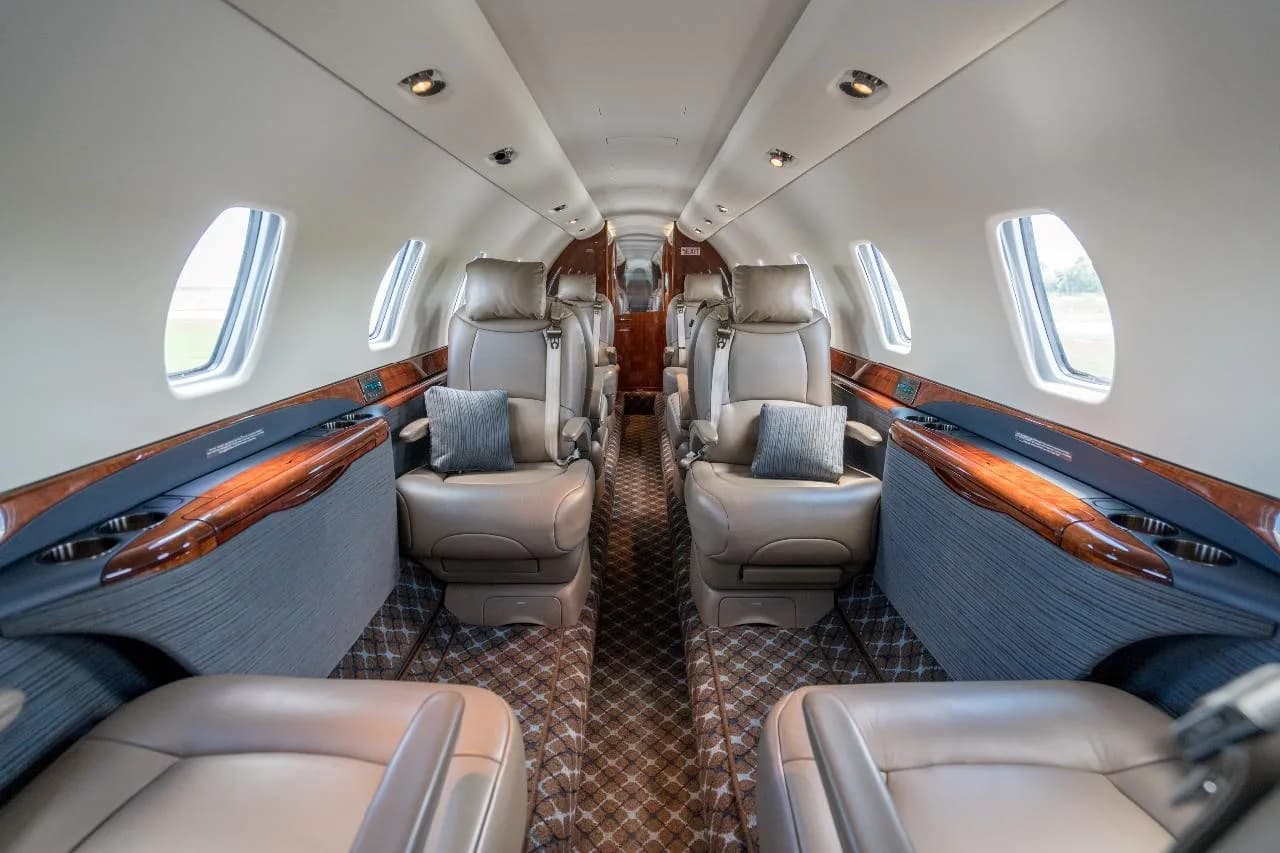 The luxurious interior cabin of a private jet features tan leather seats, wood accents, and a patterned carpet.