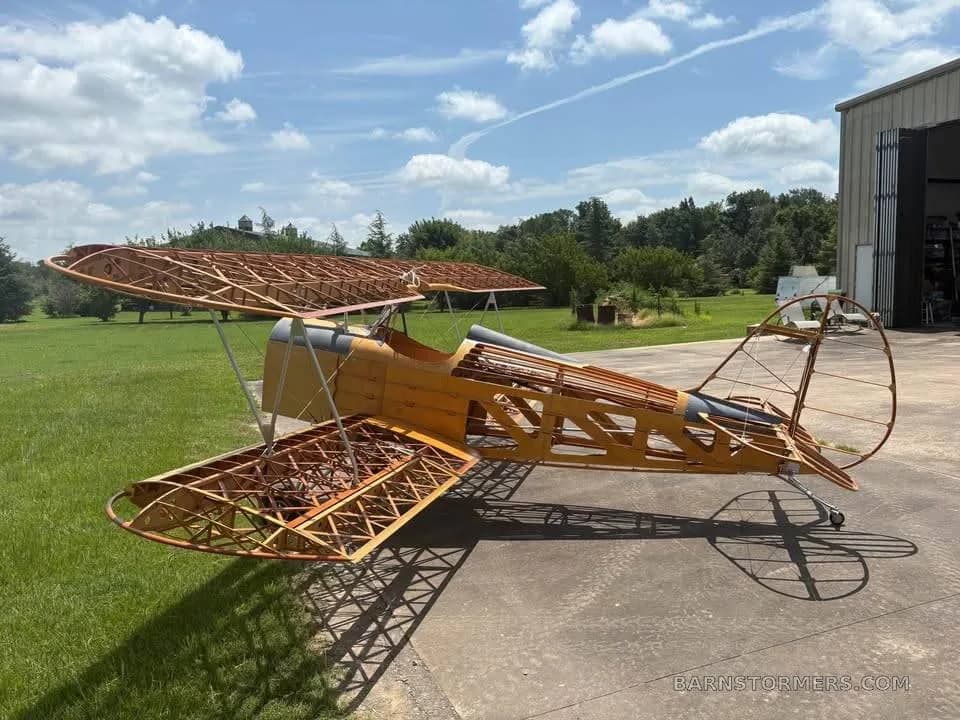 A side view of a partially assembled wooden biplane aircraft frame with exposed fuselage and wing structures, outdoors.