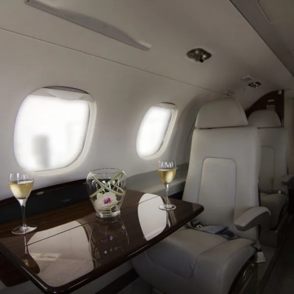 The elegant interior of a private jet, showing light grey seats, a polished wooden table with champagne, and large windows.