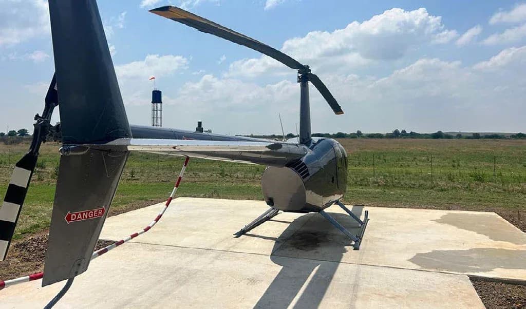 A rear-right view of a silver Robinson R44 helicopter showing the tail rotor and main rotor blades.